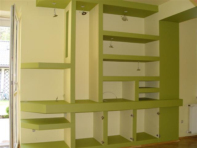 Zielone i kremowe zabudowane regały z otwartymi półkami i szafkami bez klamek, z widocznymi przewodami oświetleniowymi, w jasnym wnętrzu z oknem i grzejnikiem.